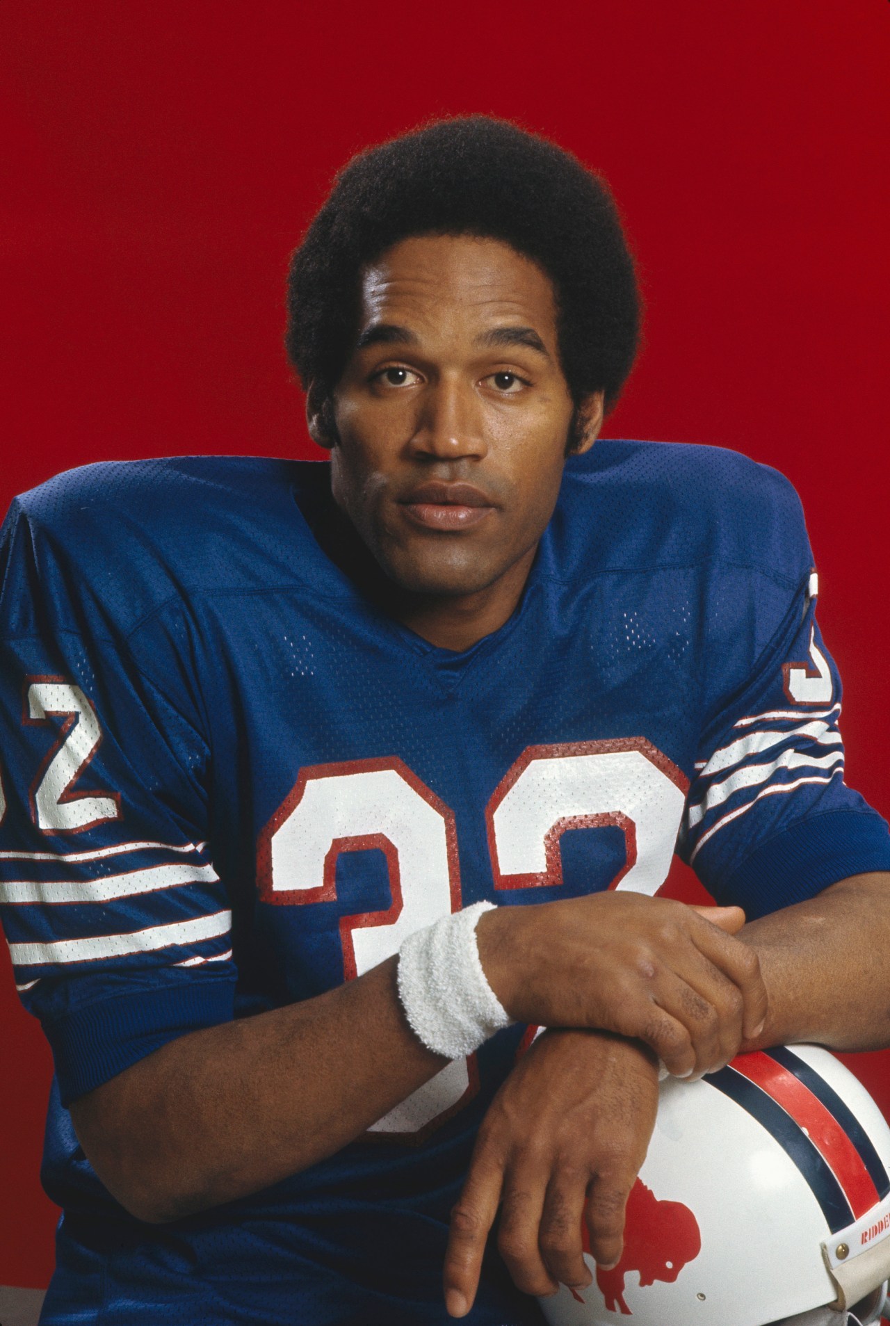 Buffalo Bills running back O.J. Simpson in blue number 32 jersey, holding a helmet, posing against a red background in the early 1970s.