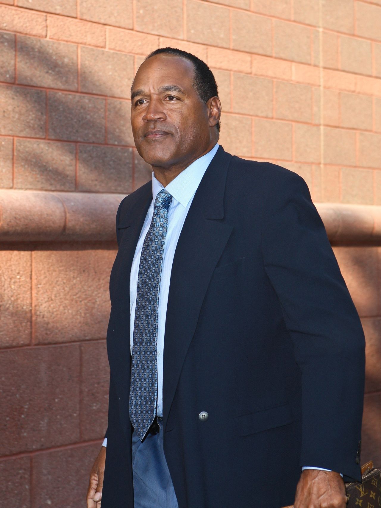 O.J. Simpson in a dark suit and blue patterned tie walking in front of a brick wall.