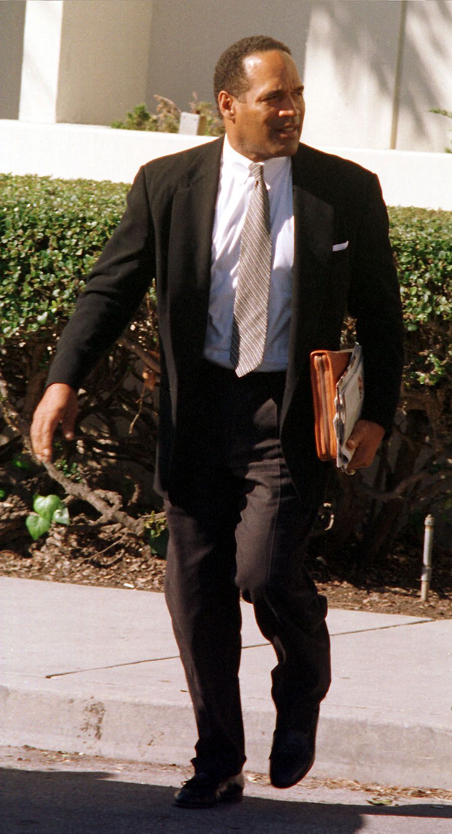O.J. Simpson in a black suit and striped tie walking along a sidewalk outside a courthouse, carrying papers.