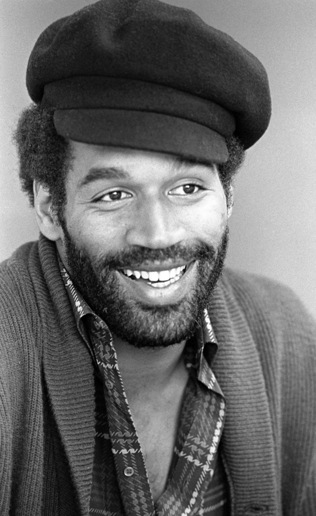 Black-and-white portrait of a smiling, bearded O.J. Simpson wearing a newsboy cap and open-collar patterned shirt.