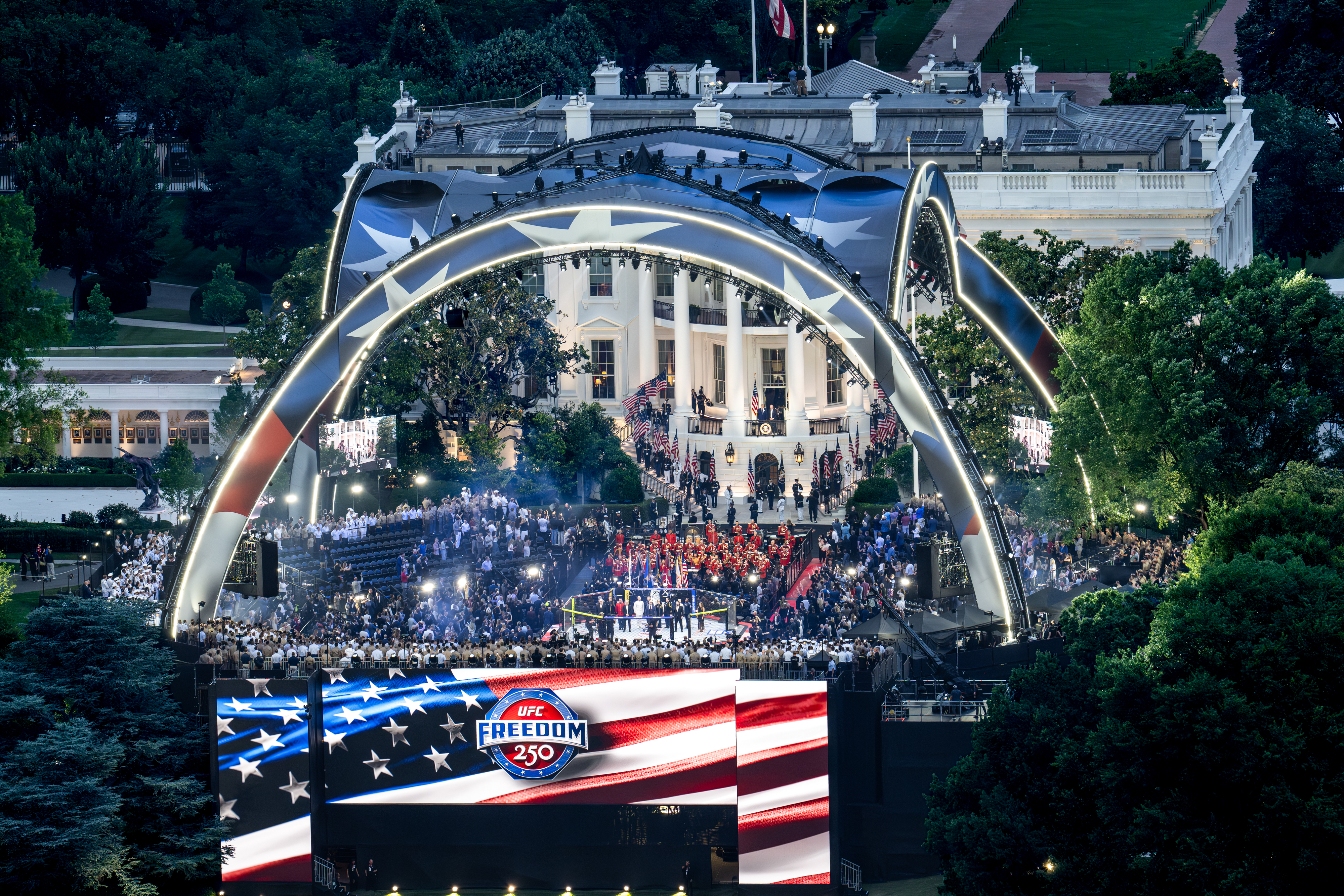 Click to open the image gallery Aerial view of UFC Freedom 250 event on the Ellipse at night, with a large illuminated arch stage, a crowd, and the White House visible behind it.