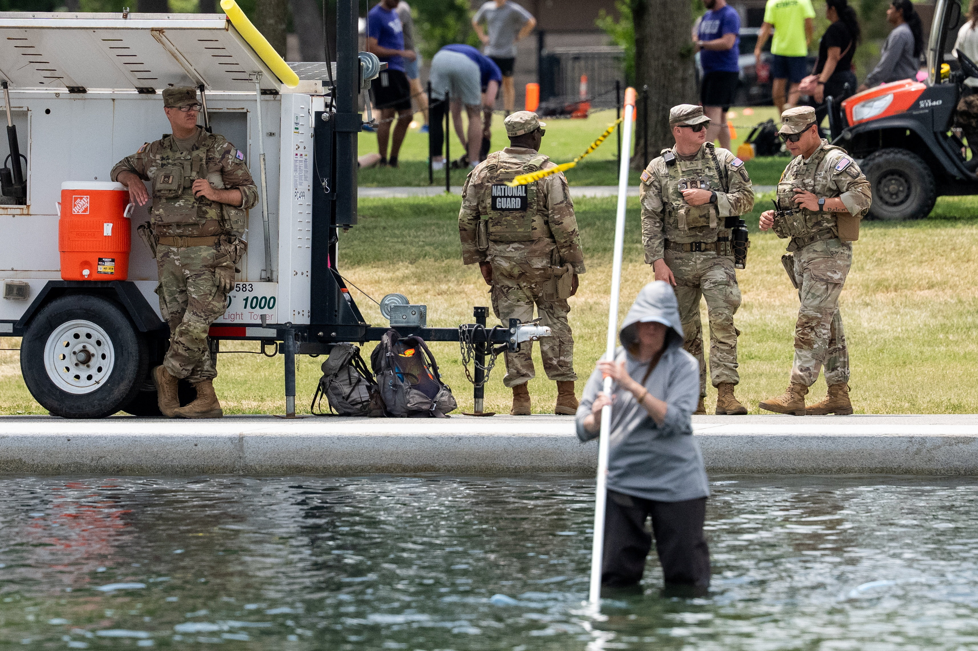 National Guard soldiers in camouflage stand near a light tower beside the Lincoln Memorial Reflecting Pool while a person wades in the water holding a pole.