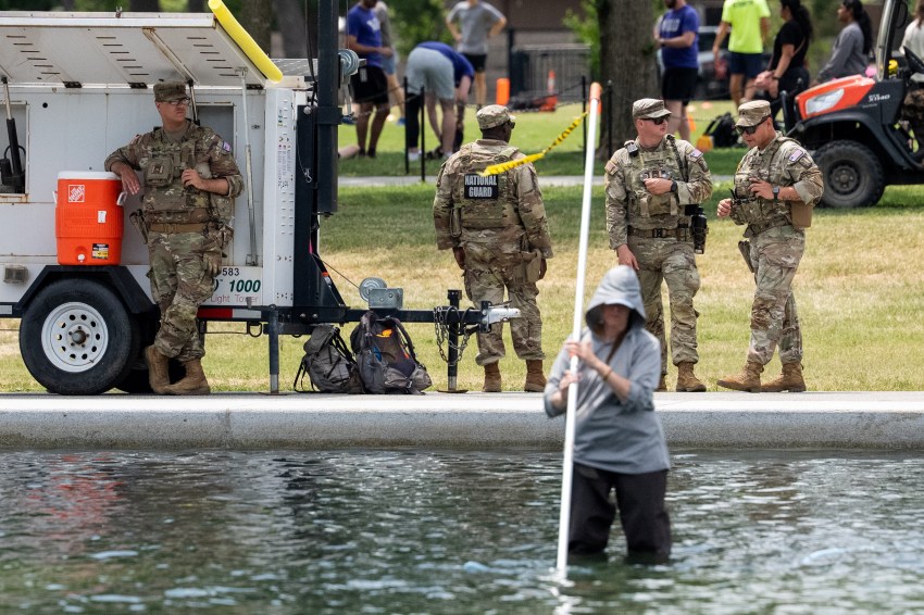 National Guard soldiers in camouflage stand near a light tower beside the Lincoln Memorial Reflecting Pool while a person wades in the water holding a pole.