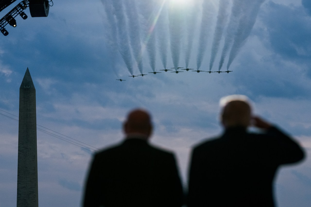 Two men seen from behind in silhouette watching a formation of jets with contrails fly over the Washington Monument.