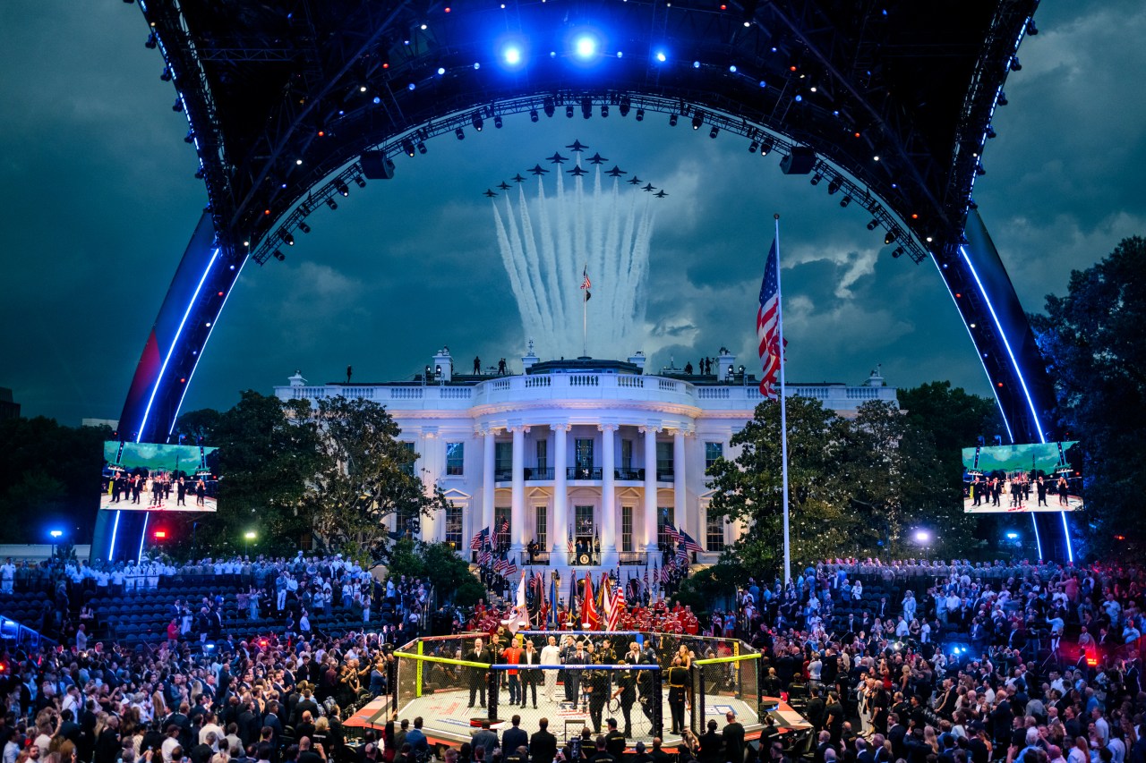 Wide aerial view of a UFC octagon and large crowd on the White House South Lawn with a jet formation trailing white smoke overhead.