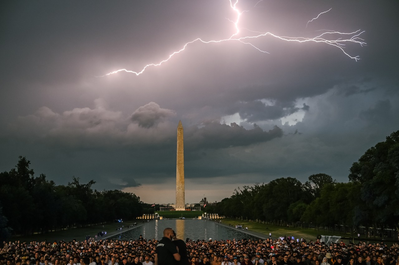 Lightning bolt striking above the Washington Monument at night with a large crowd gathered along the Lincoln Memorial Reflecting Pool in the foreground.
