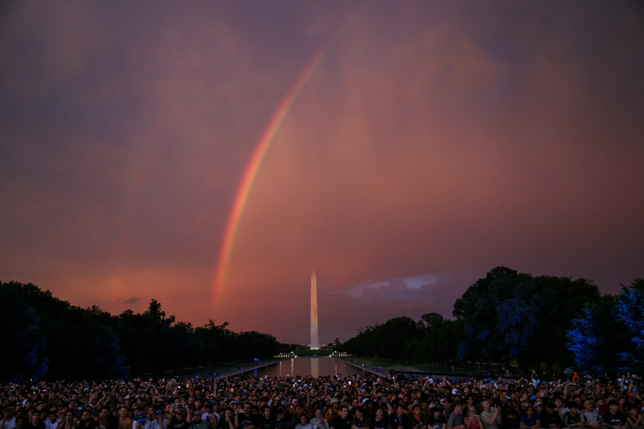 Dense crowd at the Lincoln Memorial looking toward a vivid double rainbow over the Washington Monument at sunset, reflected in the pool below.