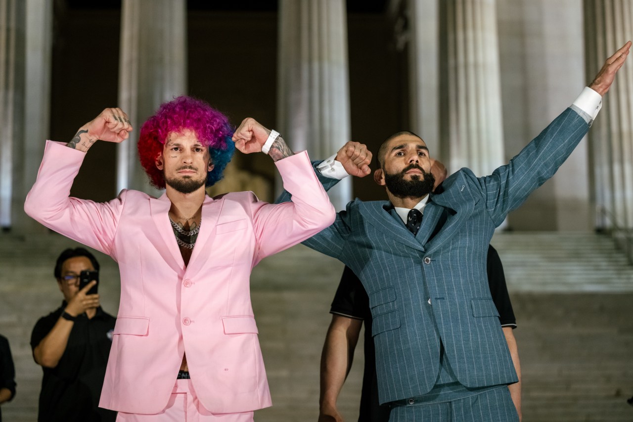 Two men posing on the steps of the Lincoln Memorial, one in a pink suit with a multicolored afro wig flexing both arms, the other in a pinstripe suit with one arm raised.