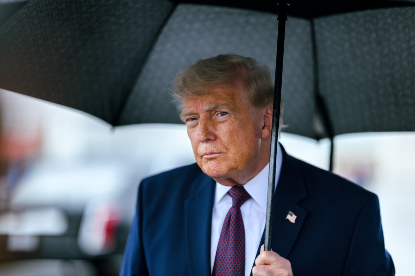 Man in blue suit and red tie holding a black umbrella at an airport tarmac, looking directly at camera.