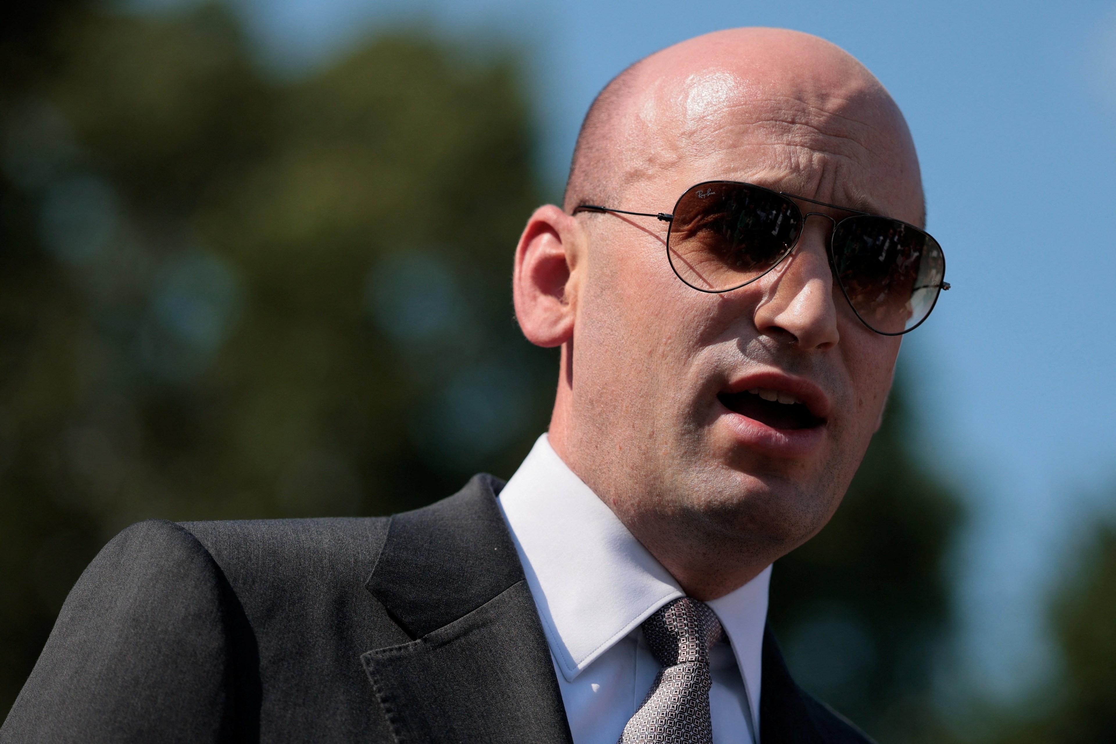 Bald man in dark suit and aviator sunglasses speaking outdoors at the White House, trees and blue sky in background.
