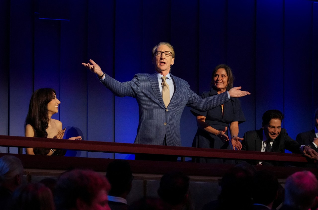 Bill Maher in a pinstripe suit stands with arms outstretched at the Kennedy Center balcony, flanked by smiling guests, during his Mark Twain Prize ceremony.