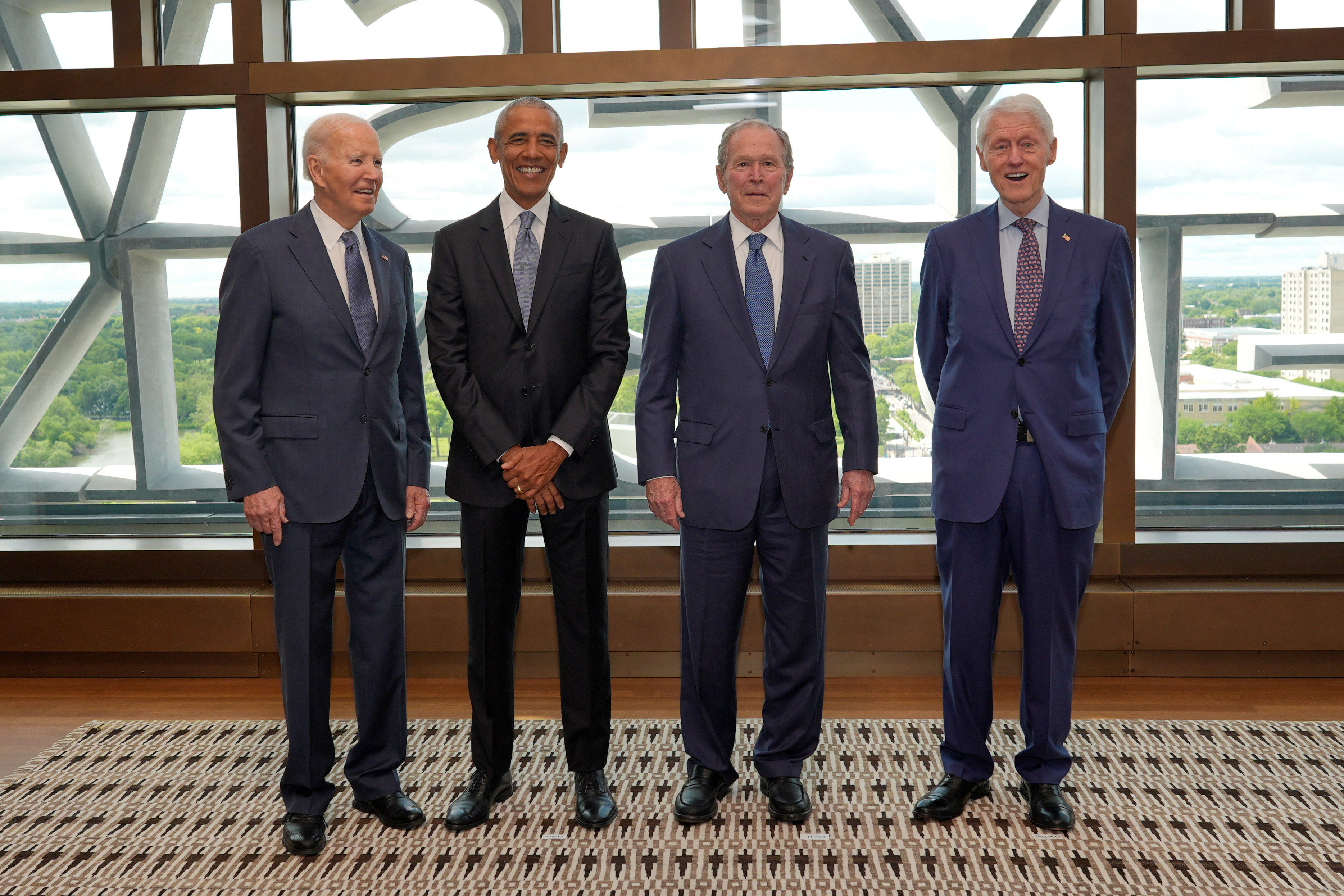 Four men in suits — Joe Biden, Barack Obama, George W. Bush, and Bill Clinton — pose together in front of large windows overlooking a cityscape.