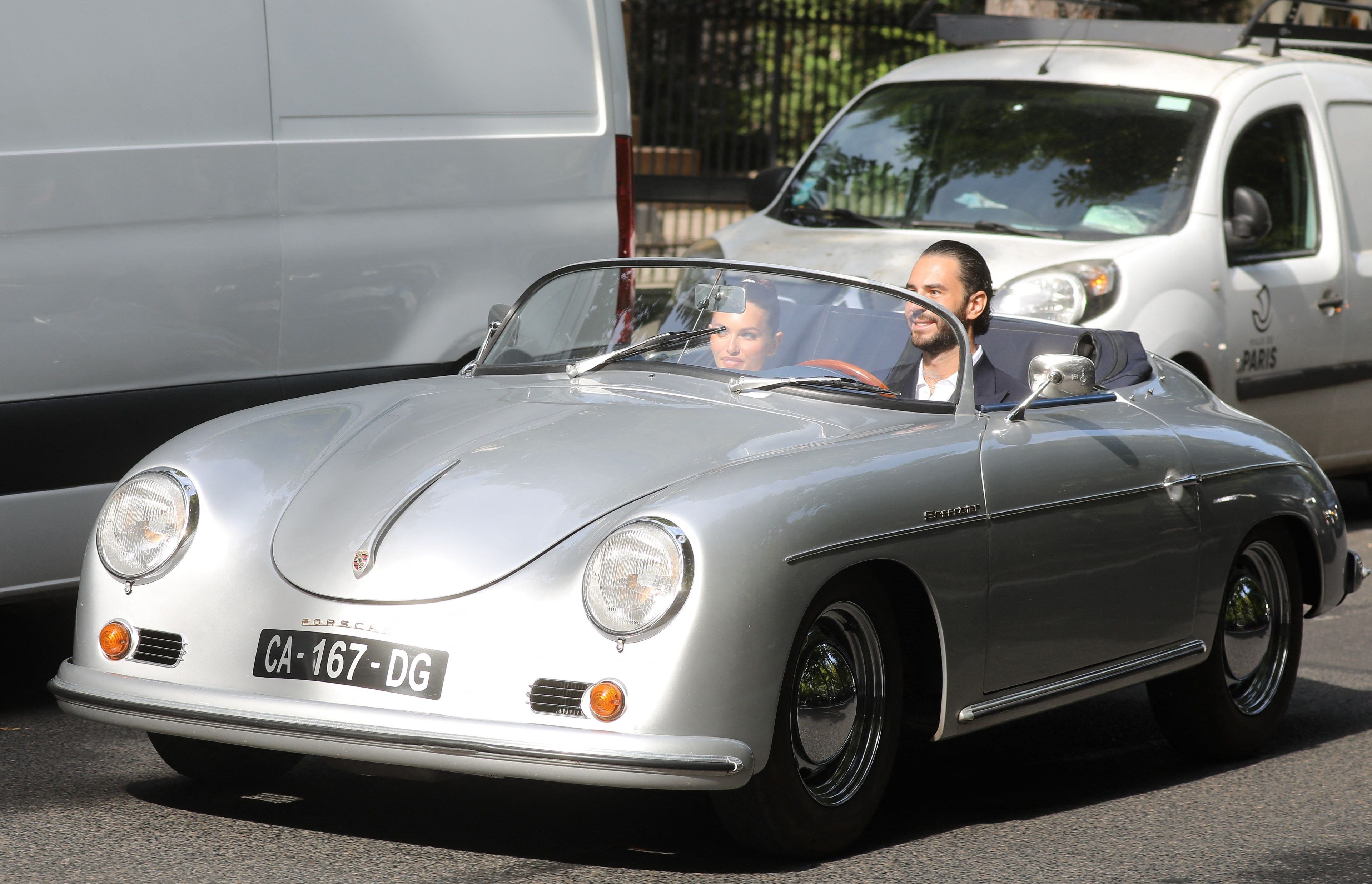 Thylane Blondeau and Benjamin Attal riding in a vintage silver Porsche 356 Speedster convertible on a Paris street.