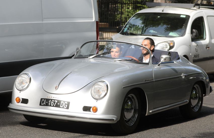 Thylane Blondeau and Benjamin Attal riding in a vintage silver Porsche 356 Speedster convertible on a Paris street.