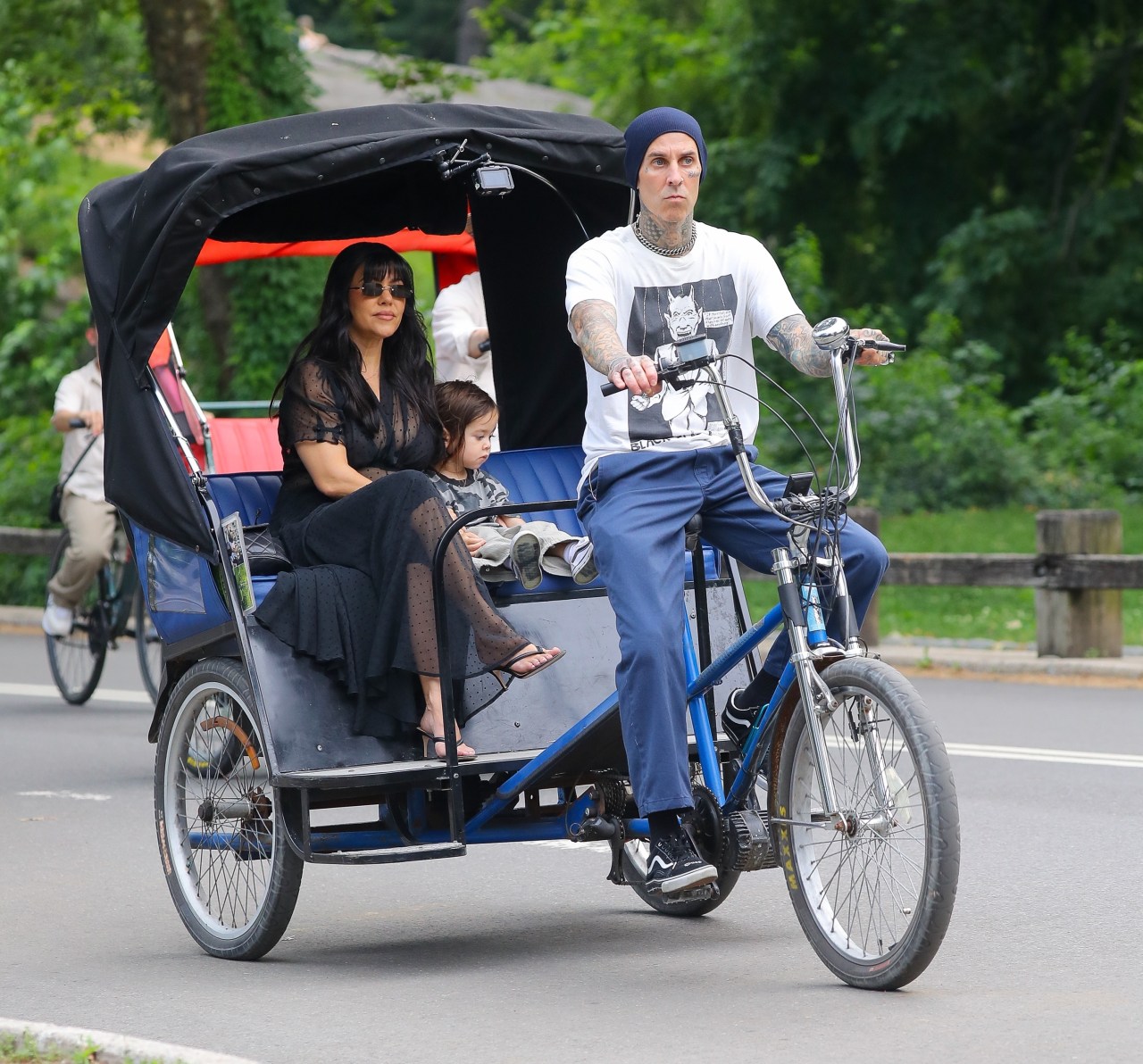Tattooed man in white t-shirt and blue beanie pedaling a blue pedicab, with a dark-haired woman in black and a young child seated behind him in Central Park.