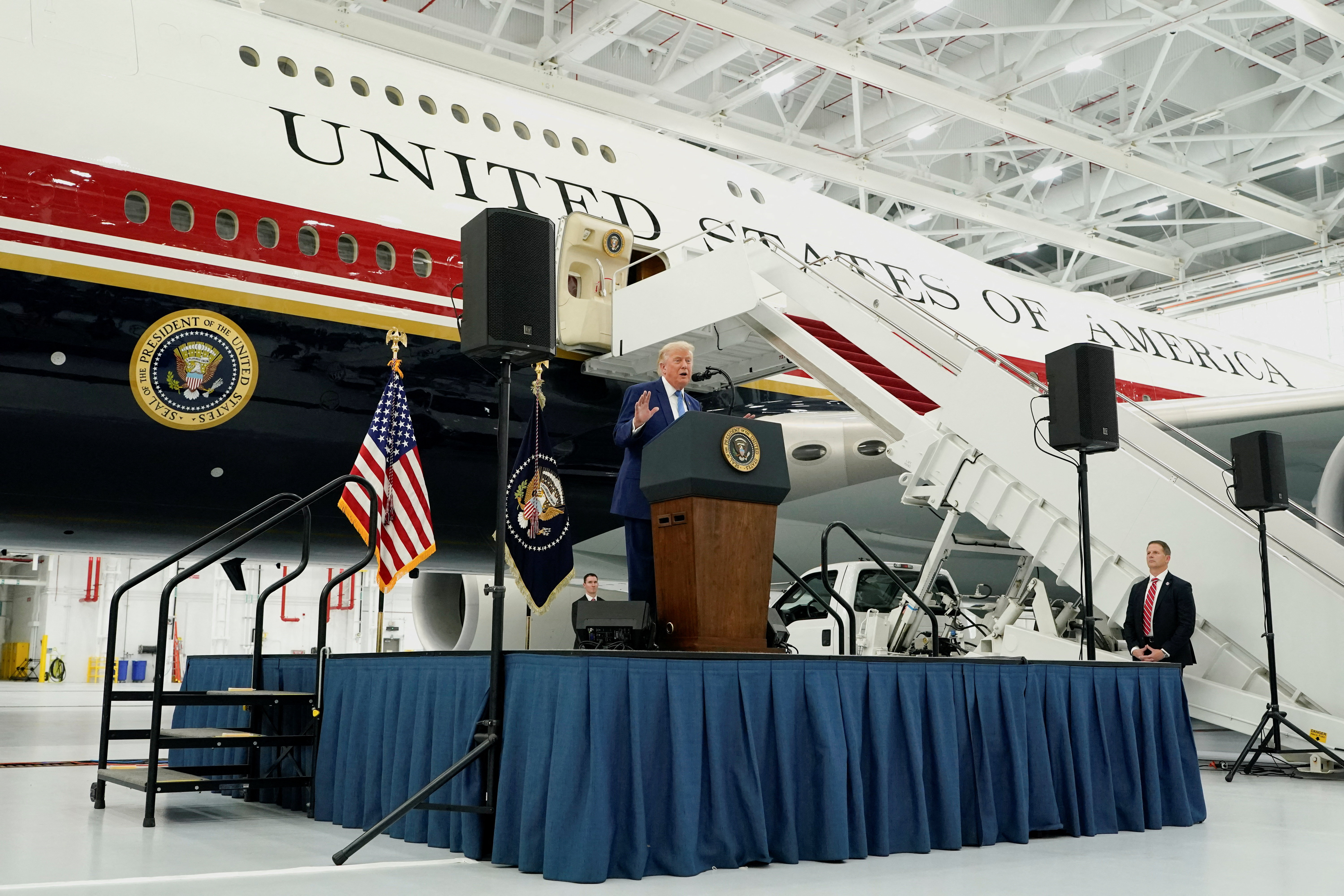 Click to open the image gallery A man in a blue suit speaks at a presidential podium in front of a large white jet marked 'United States of America' with its boarding stairs deployed inside a hangar.