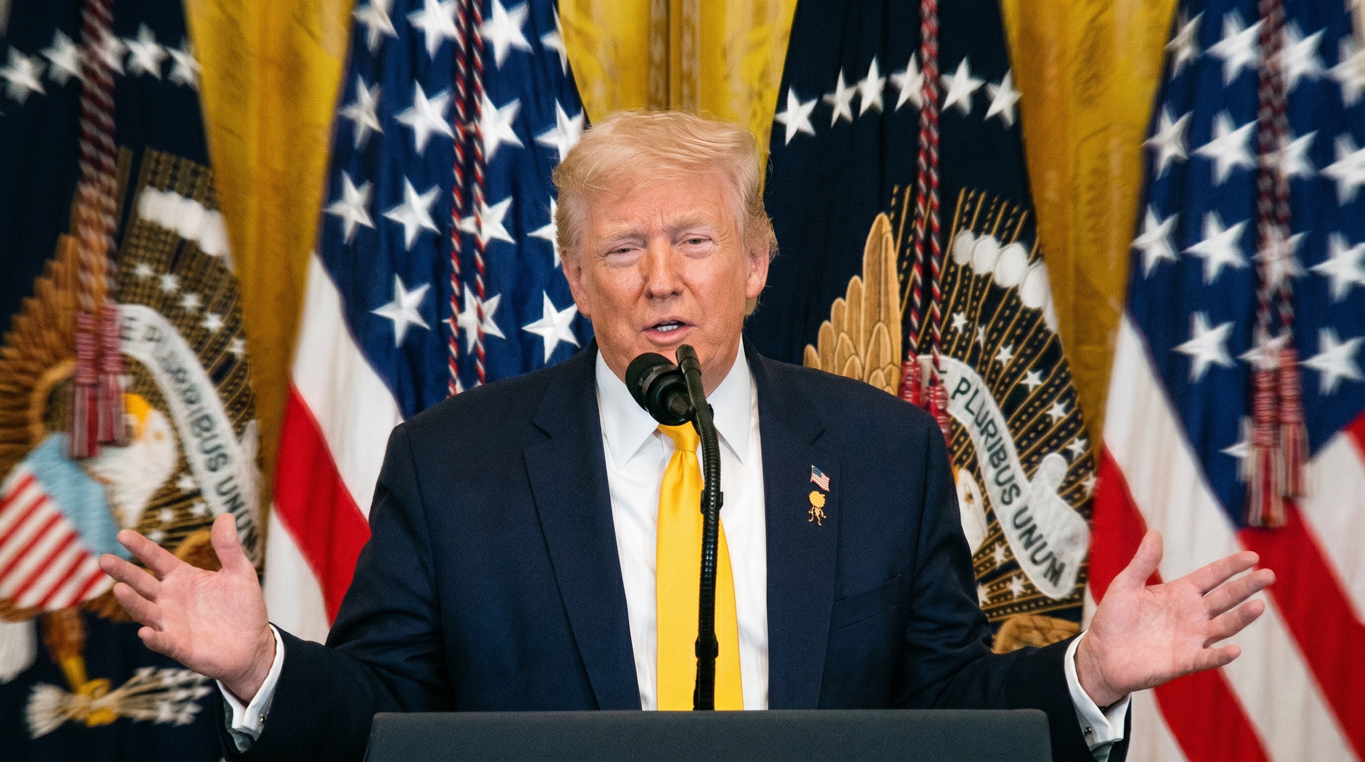 Man in navy suit and yellow tie speaking at a podium with American flags and Presidential Seal banners in the background.
