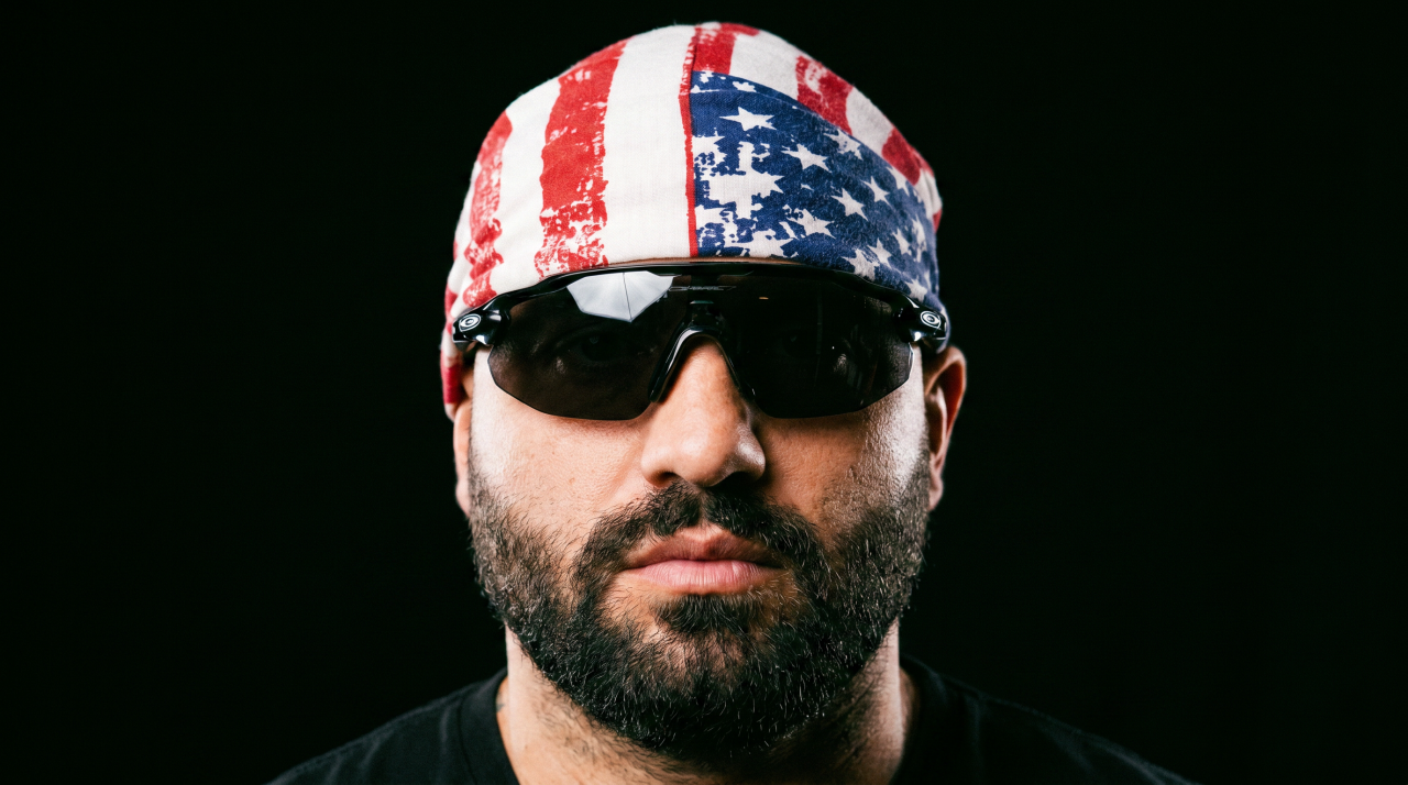 Bearded man wearing an American flag do-rag and dark wraparound sunglasses against a black background.