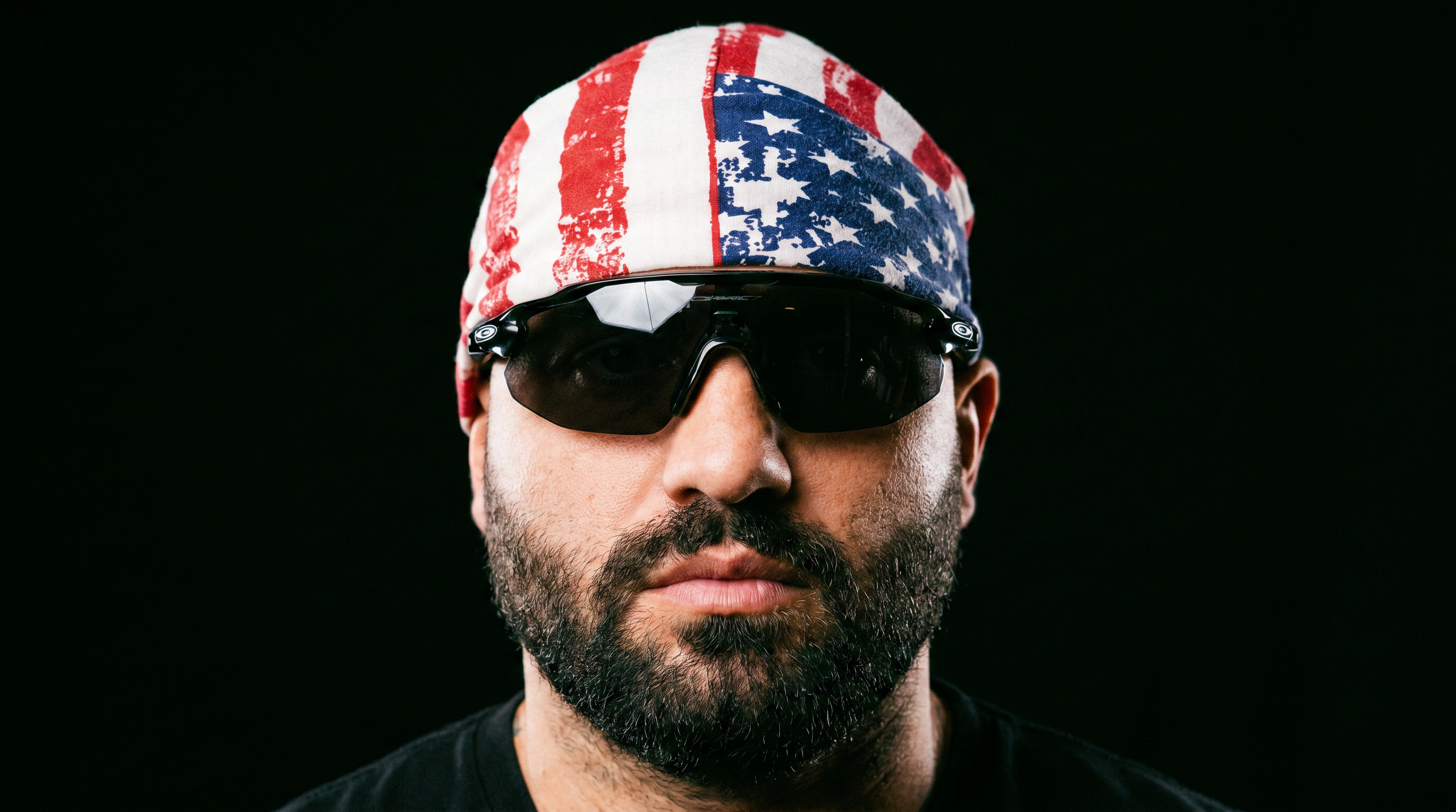 Bearded man wearing an American flag do-rag and dark wraparound sunglasses against a black background.