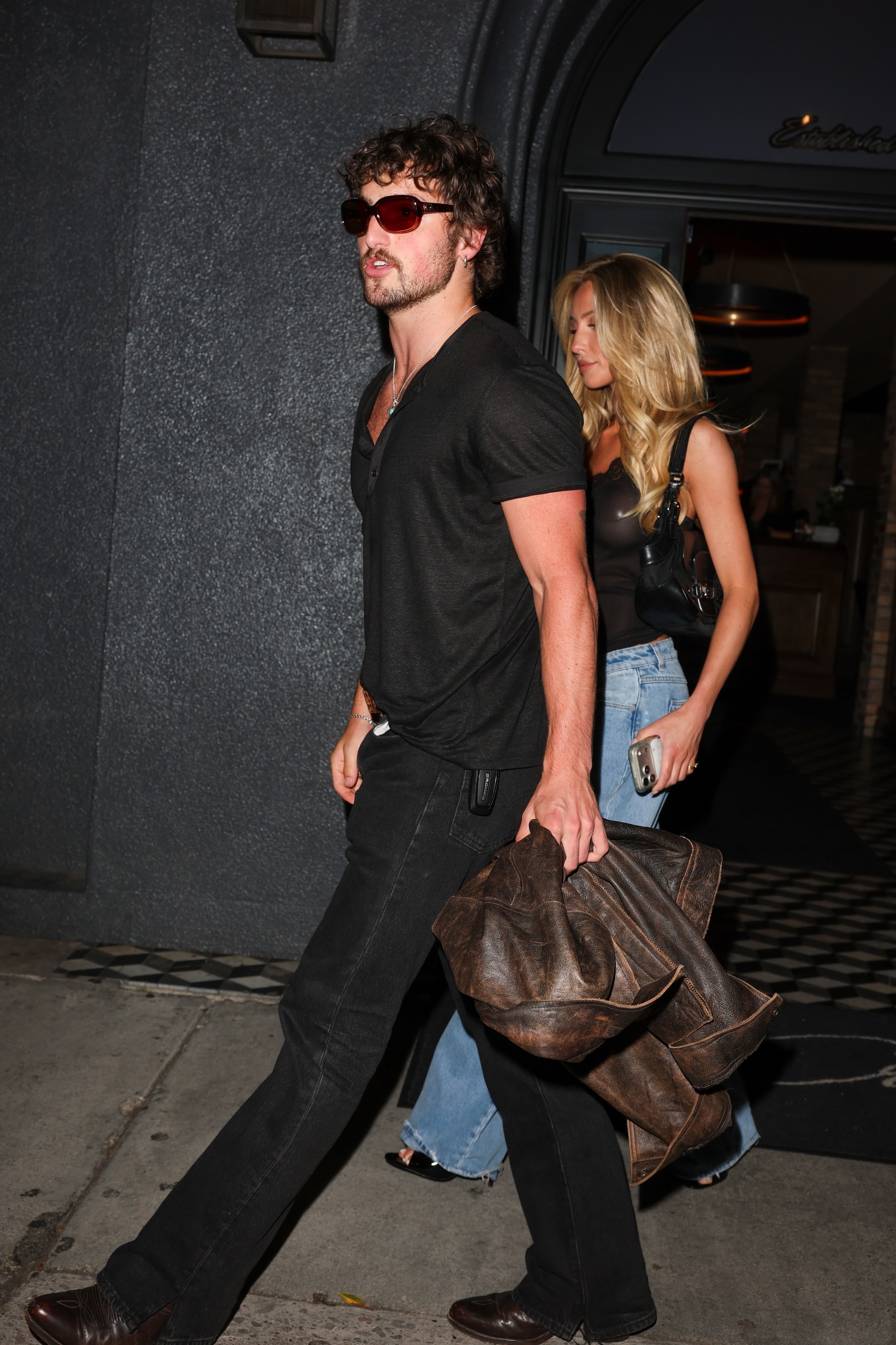 Click to open the image gallery A curly-haired man in a black t-shirt and dark jeans carries a large brown leather bag while a blonde woman in blue jeans walks behind him outside a building at night.