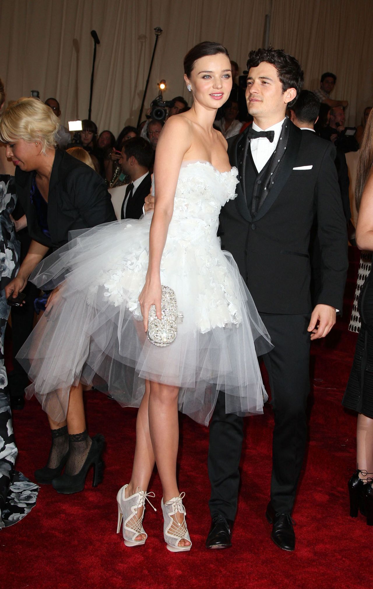 Woman in white strapless tulle mini gown holding a crystal clutch stands beside a man in a black tuxedo on a red carpet.