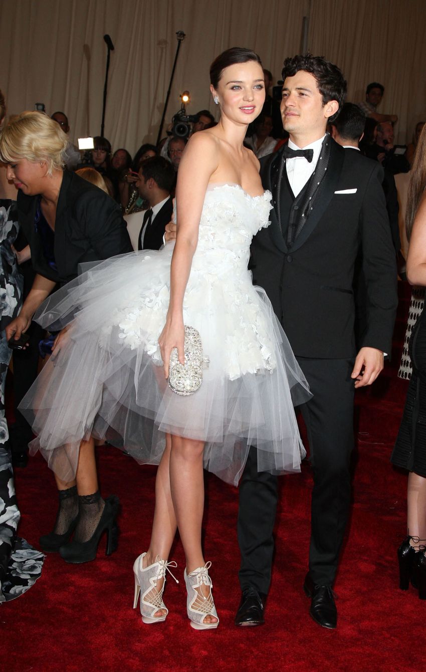 Woman in white strapless tulle mini gown holding a crystal clutch stands beside a man in a black tuxedo on a red carpet.