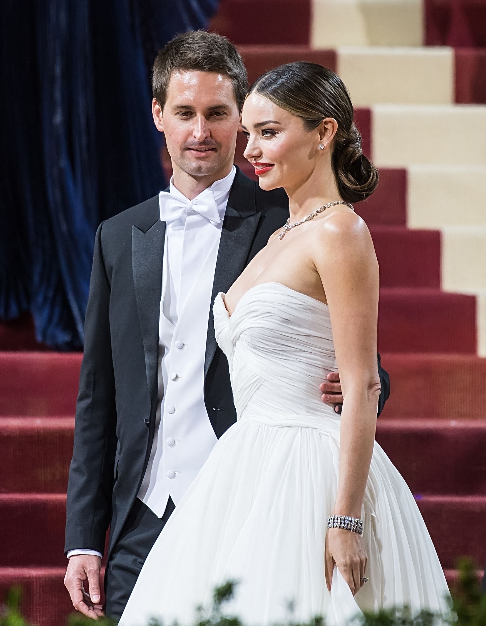 Click to open the image gallery Woman in strapless white draped gown and man in black tailcoat with white bow tie pose together on ornate red and cream stairs.