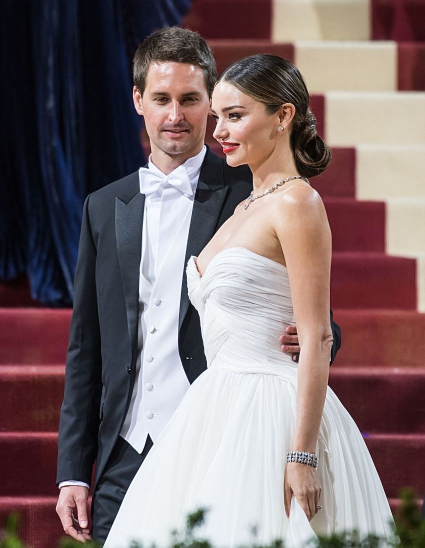 Woman in strapless white draped gown and man in black tailcoat with white bow tie pose together on ornate red and cream stairs.