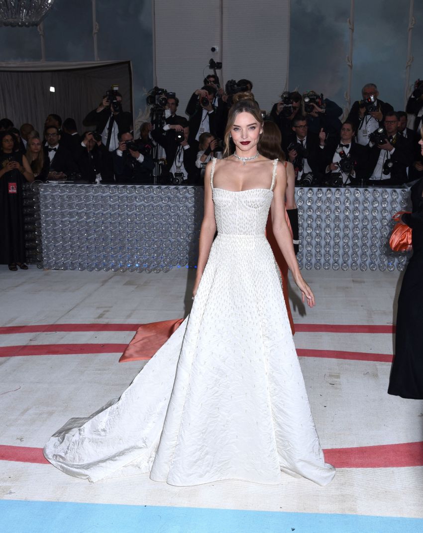 Woman in a white beaded strapless ball gown with a long train poses at a gala inside a grand decorated hall with photographers behind her.