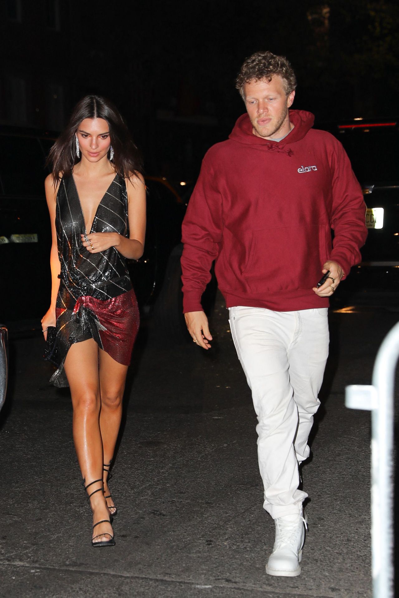 A brunette woman in a deep-V metallic wrap minidress and strappy sandals walks alongside a man in a red hoodie and white jeans at night.