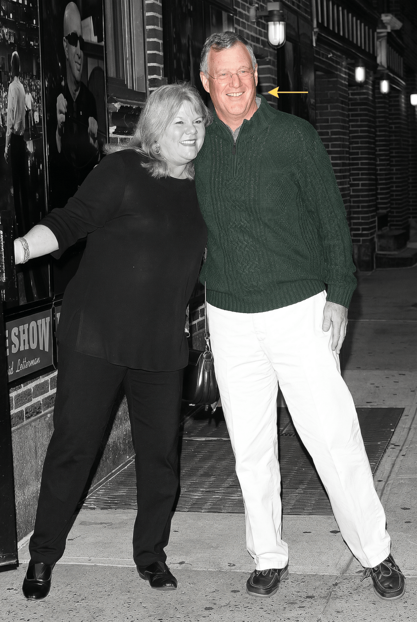 A man in a green sweater and white pants and a woman in all black smile together outside the Late Show with David Letterman, in a black-and-white photo with the man's sweater colorized green.