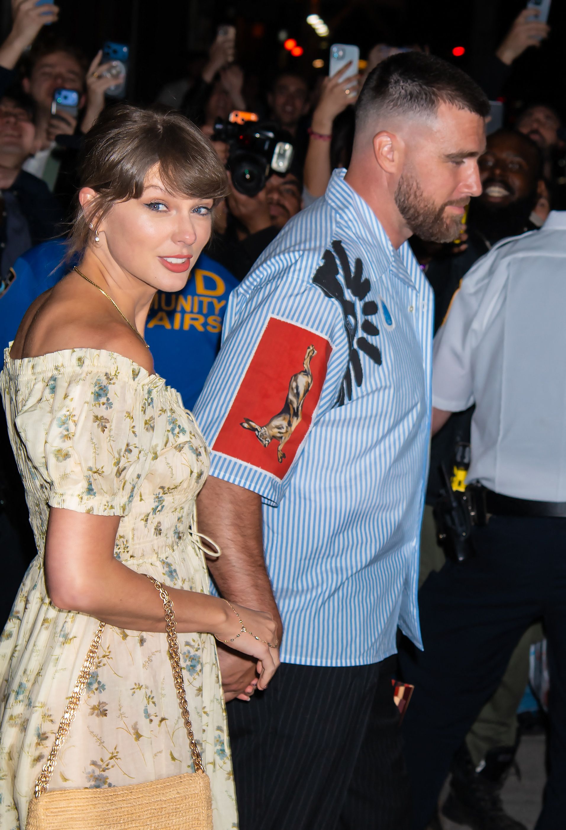 Click to open the image gallery A woman in a cream floral off-shoulder dress and a tall bearded man in a blue striped shirt walk hand-in-hand through a crowd of fans and photographers at night.