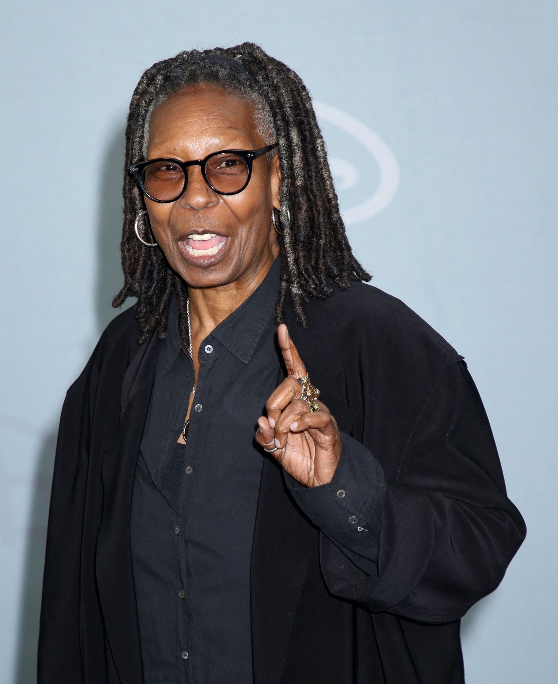 Whoopi Goldberg in all black with dreadlocks and round glasses, pointing one finger upward and speaking at the 2026 Disney Upfront event.
