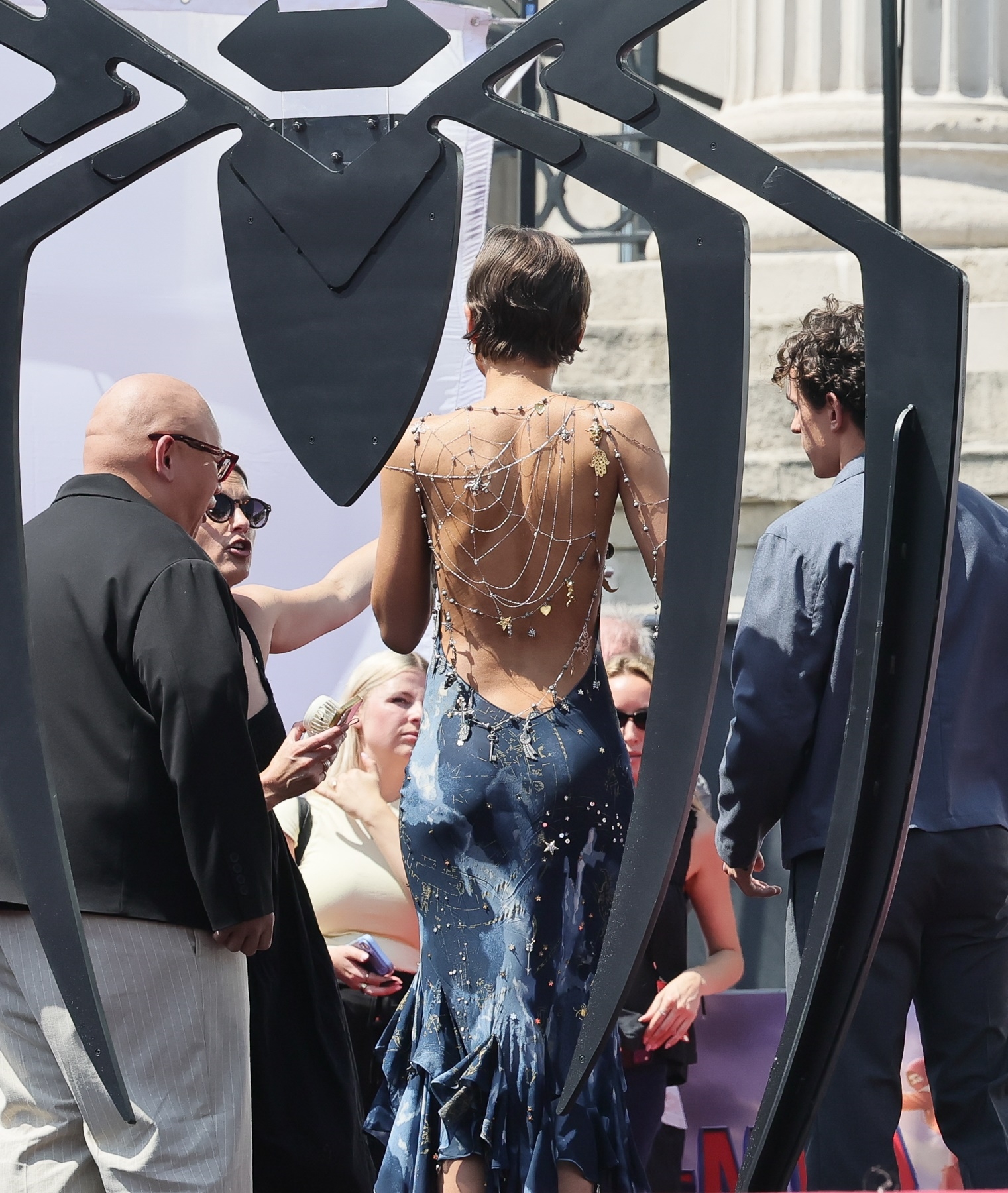 Click to open the image gallery Back view of woman in backless blue dress with intricate silver spider web chain body jewelry covering her entire back, standing near a large black spider-shaped structure.