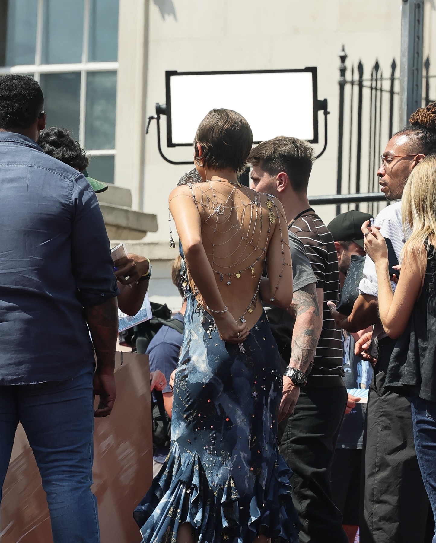 Click to open the image gallery Back view of woman in backless blue ruffle-hem dress with silver spider web chain jewelry draped across her back, standing amid a crowd on a red carpet.