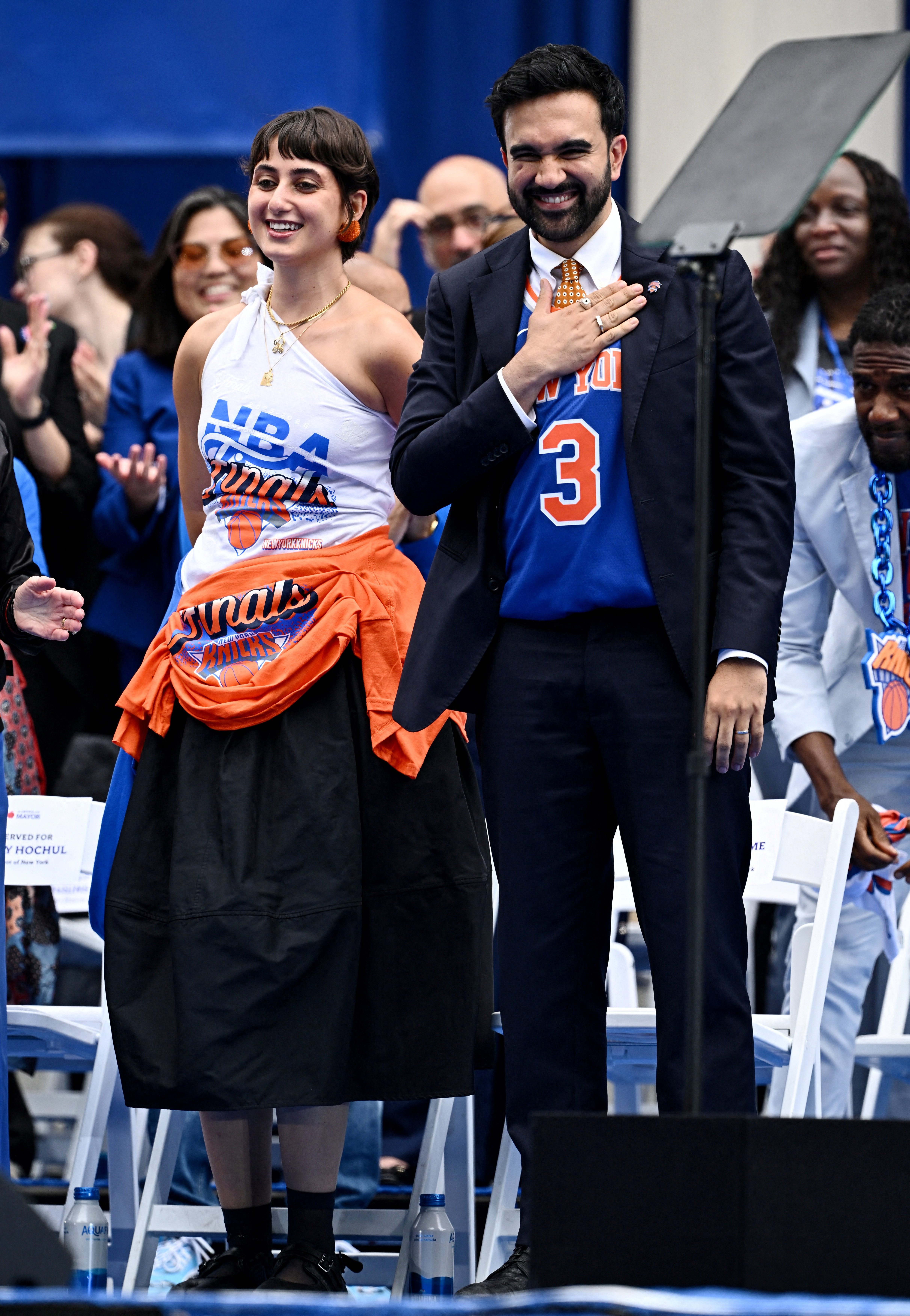 Zohran Mamdani in a suit and Knicks jersey smiling with Rama Duwaji in an NBA Finals top and orange shirt tied at her waist, standing at NYC City Hall.