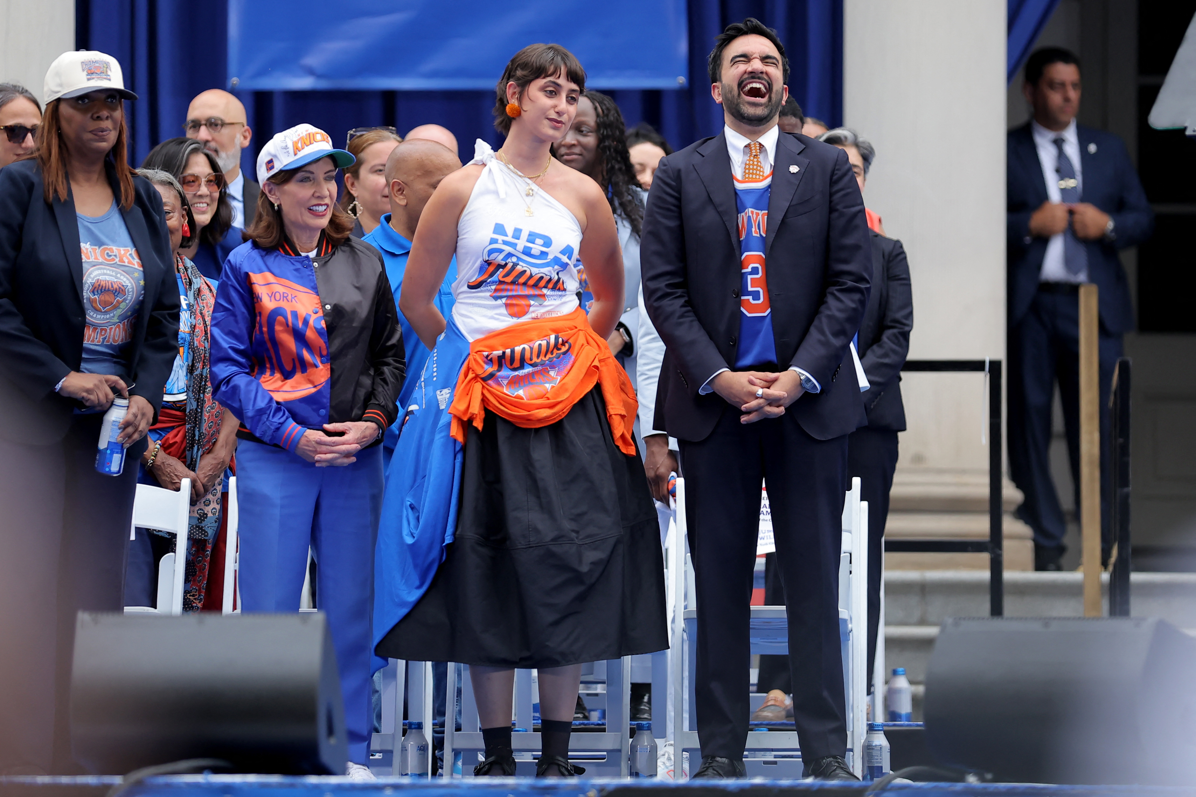 Zohran Mamdani laughing loudly beside Rama Duwaji and Kathy Hochul in a Knicks jacket at the NYC City Hall championship ceremony.
