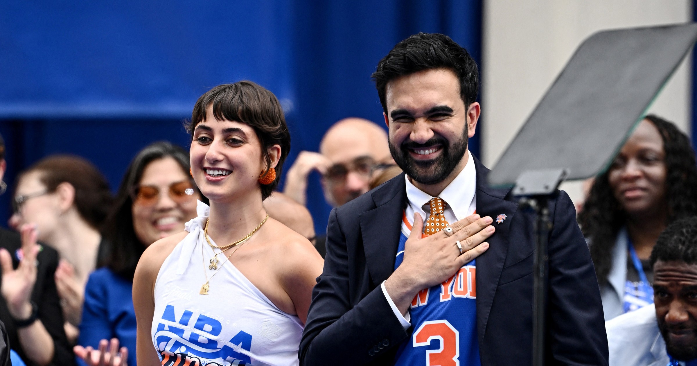 Zohran Mamdani in a navy suit with a blue Knicks jersey over it stands smiling next to Rama Duwaji, who wears a white NBA Finals tank top and black skirt with an orange Knicks shirt tied at the waist.