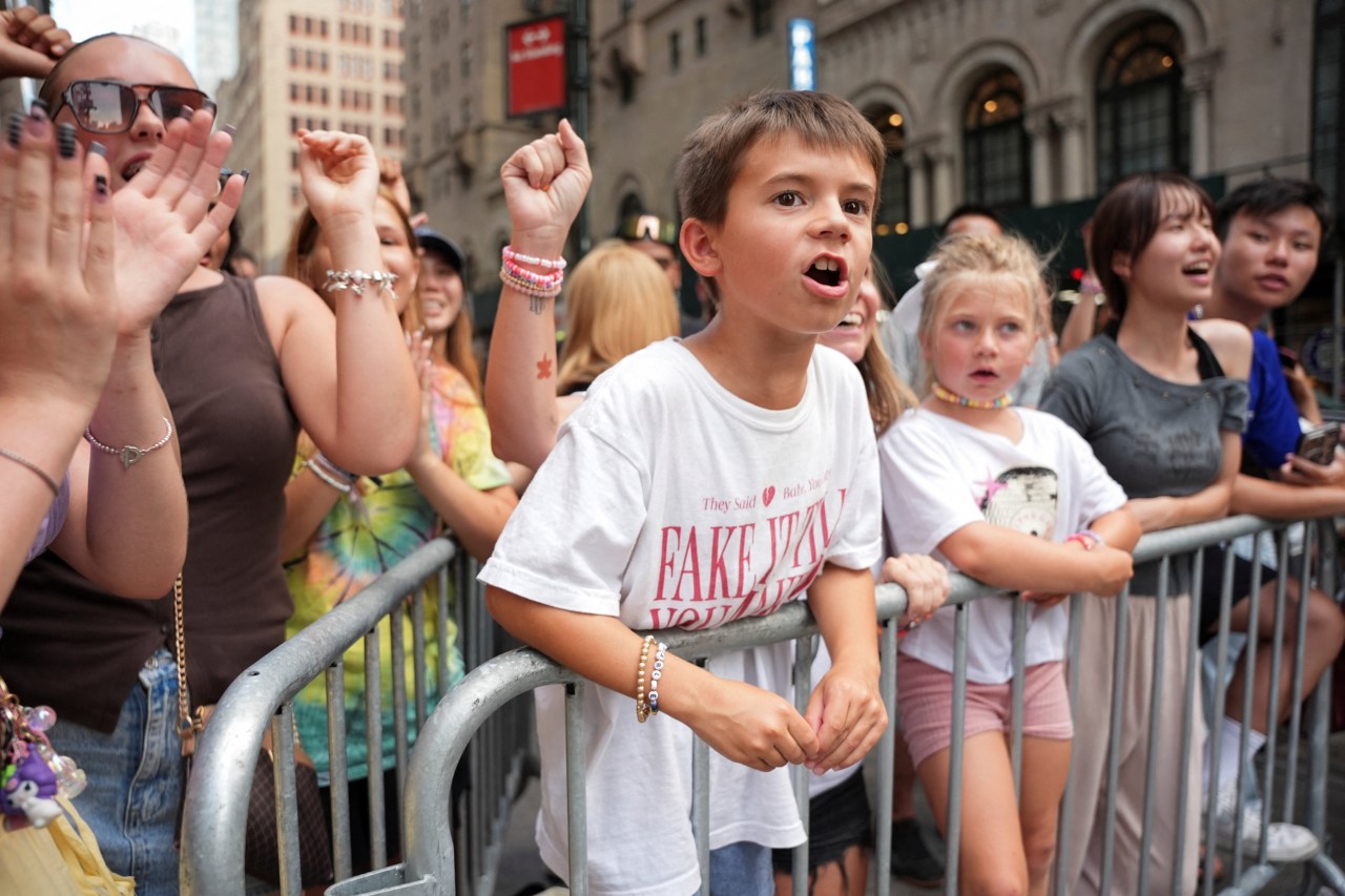 Excited young boy in a white Taylor Swift T-shirt raises his fist at a crowd barrier outside Madison Square Garden, surrounded by other fans.