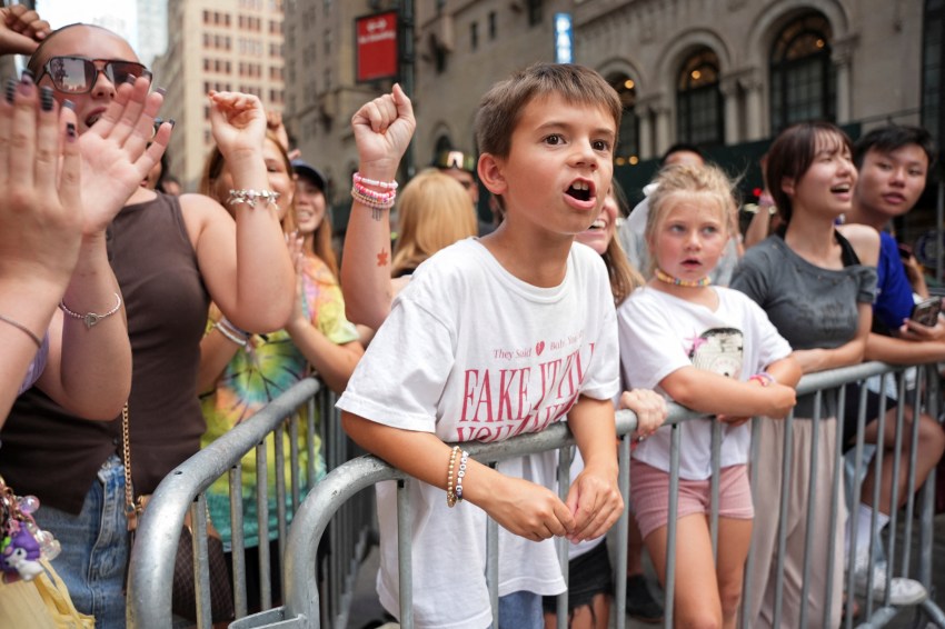 Excited young boy in a white Taylor Swift T-shirt raises his fist at a crowd barrier outside Madison Square Garden, surrounded by other fans.