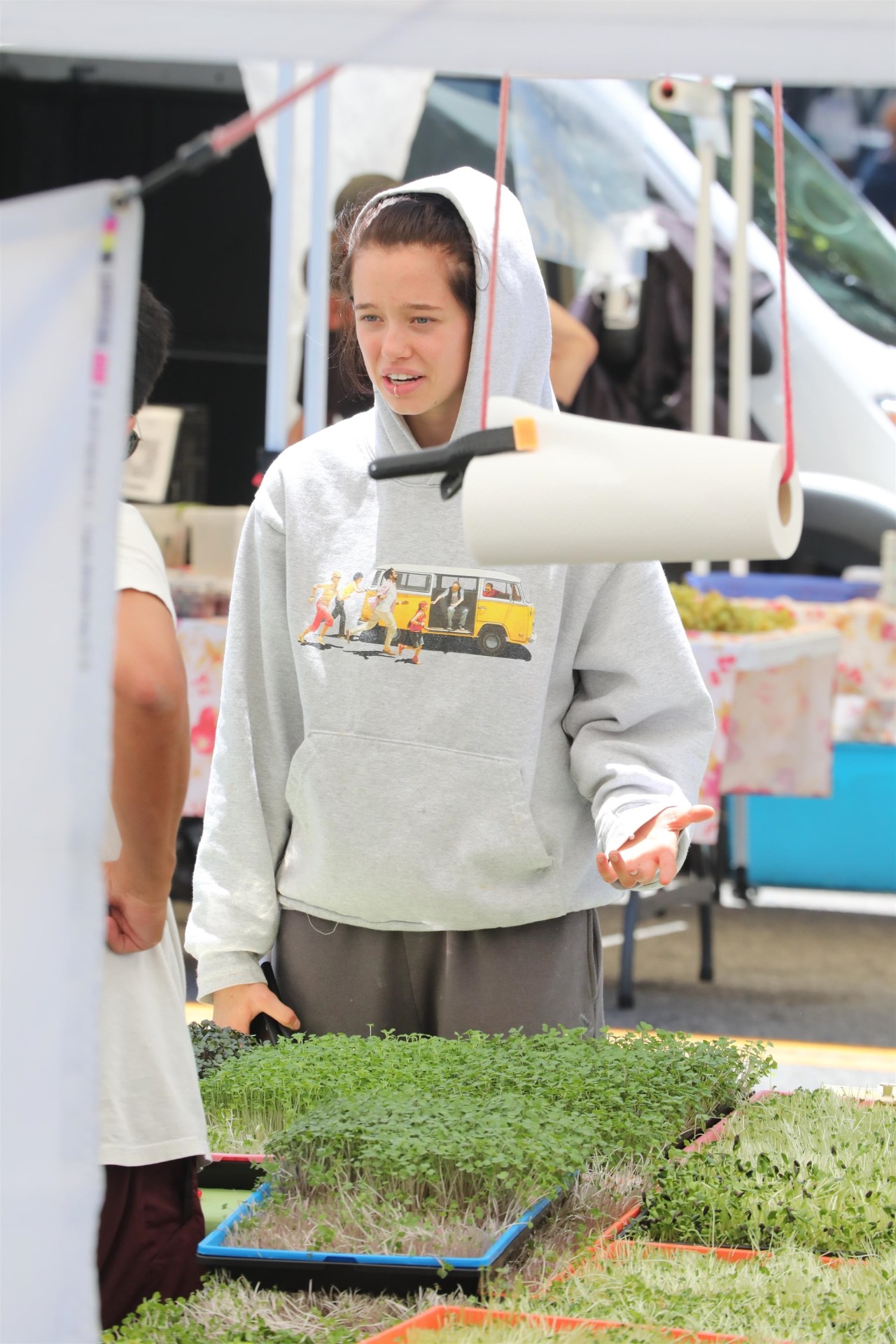 Teenage girl in a gray hoodie leaning over trays of microgreens at an outdoor farmers market stall.