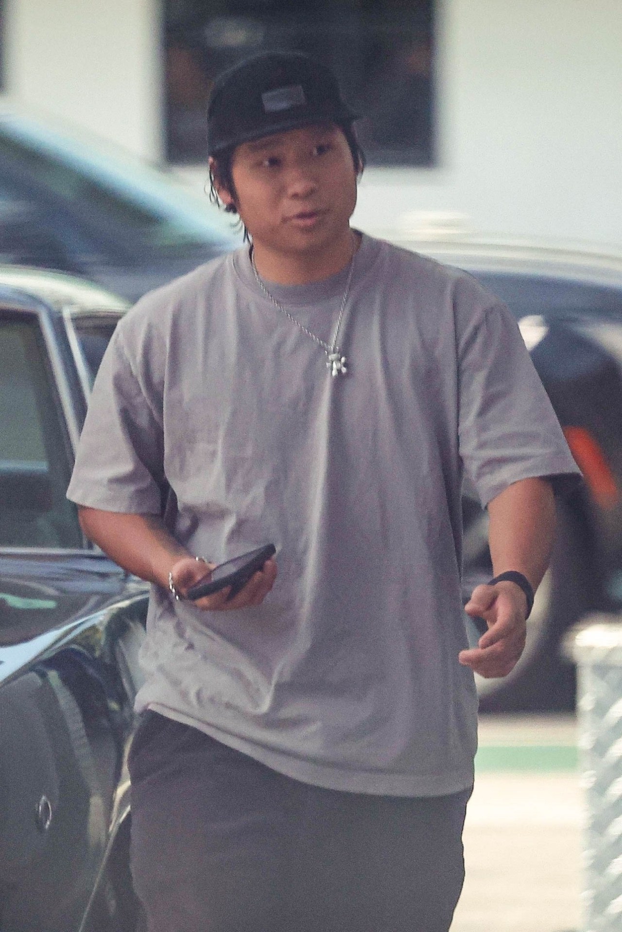 Young man in a gray t-shirt and black cap holding a smartphone while walking near a dark car in a parking area.
