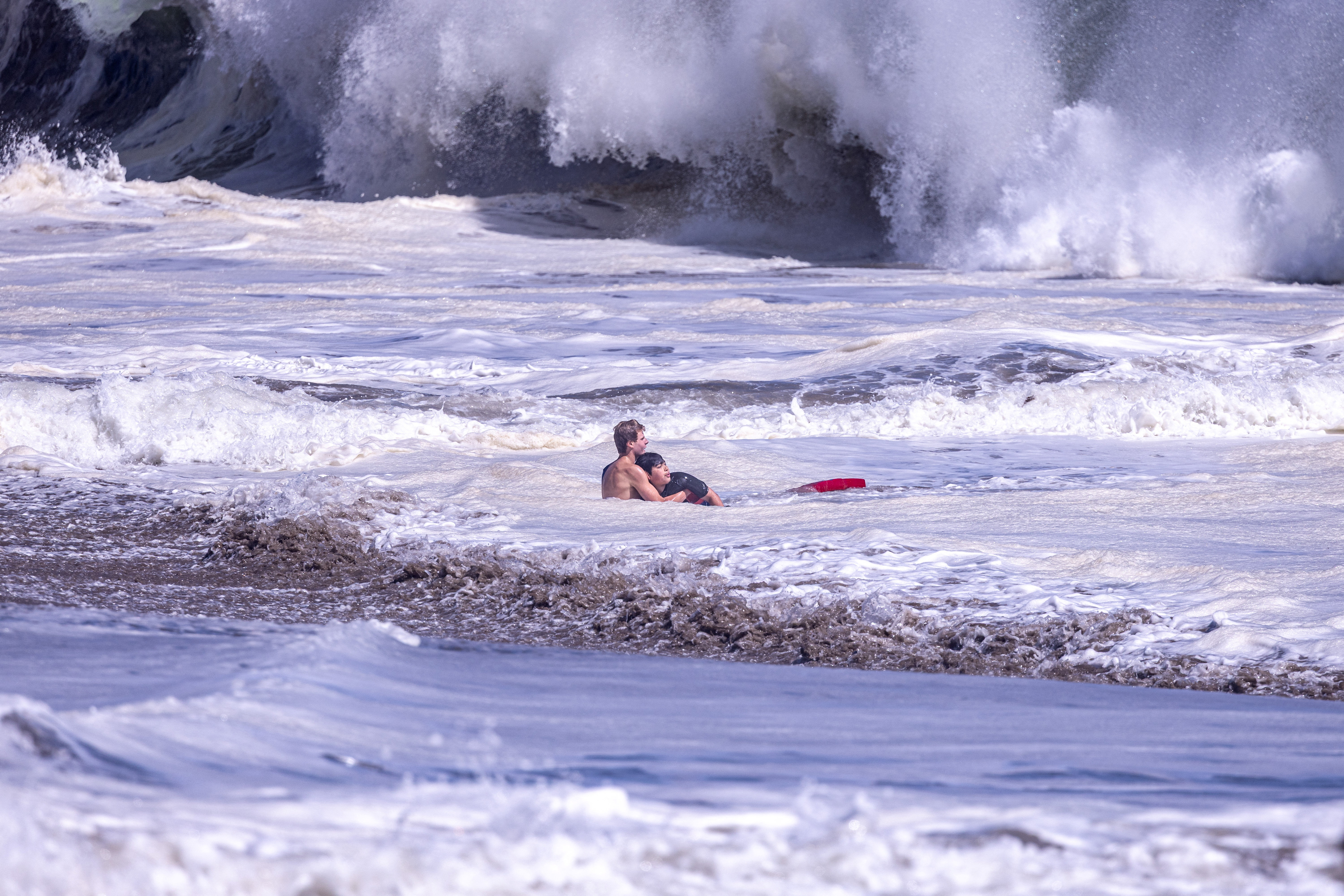 A shirtless lifeguard holds a young boy in turbulent, foamy ocean surf as enormous waves crash behind them at Seabright Beach.