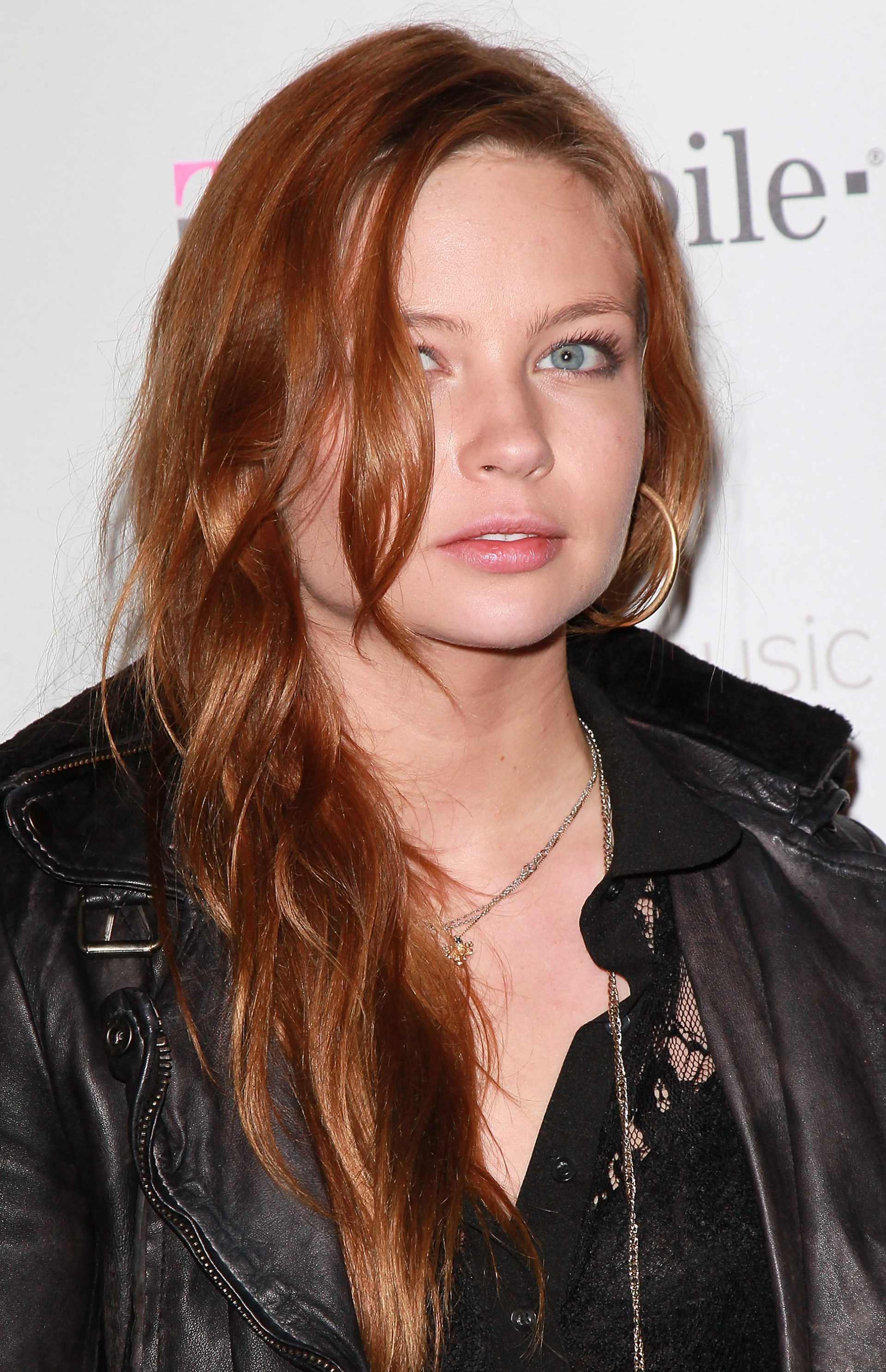 Daveigh Chase with long auburn hair wearing a black leather jacket and lace top at a T-Mobile event backdrop.
