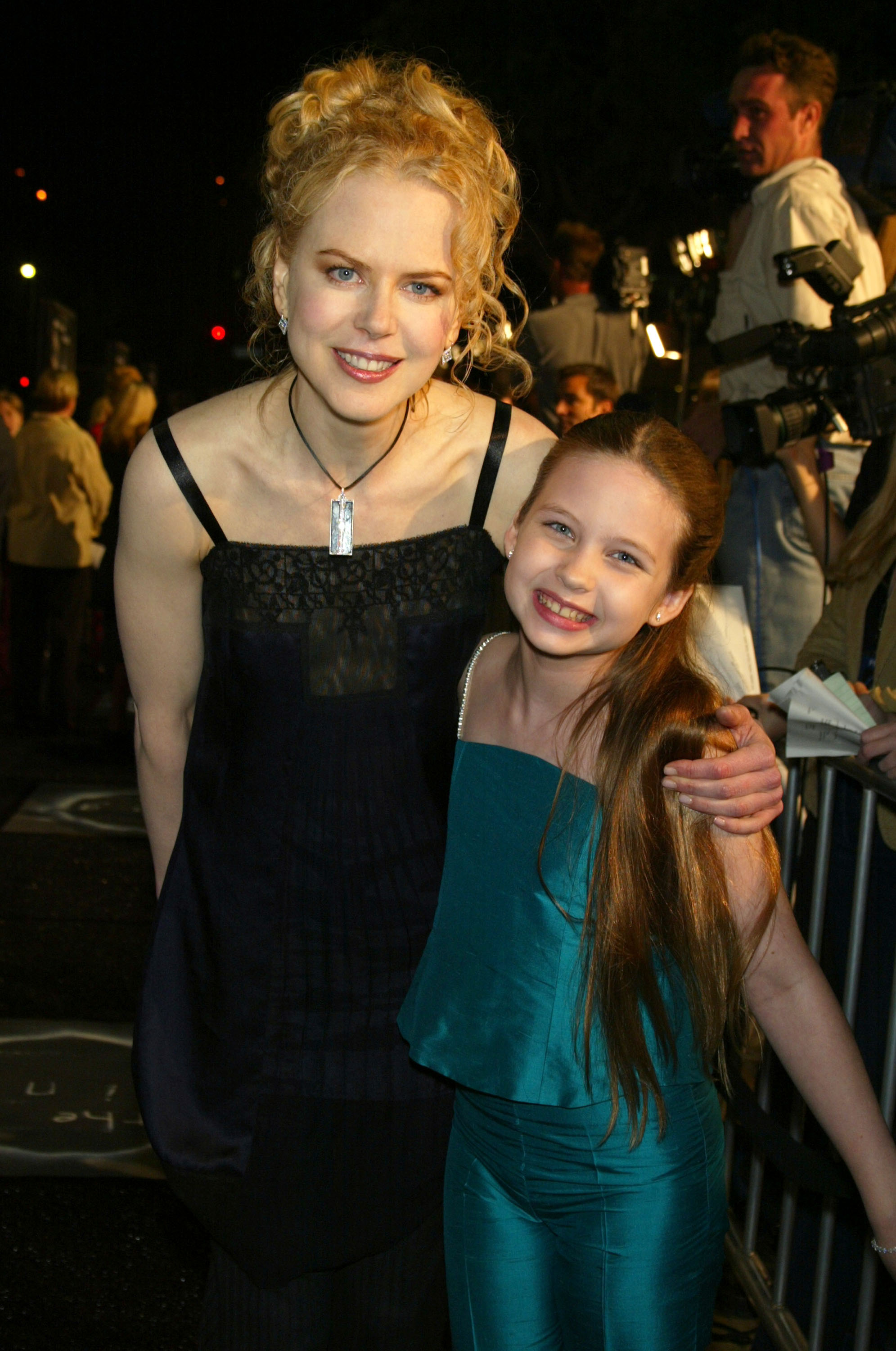 Click to open the image gallery A woman in a black dress and a young red-haired girl in a teal outfit smile together at a movie premiere, with cameras visible in the background.