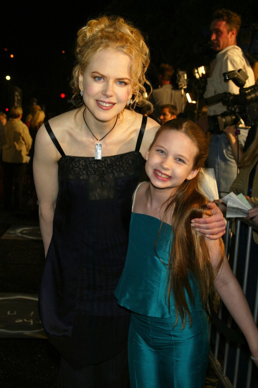 A woman in a black dress and a young red-haired girl in a teal outfit smile together at a movie premiere, with cameras visible in the background.