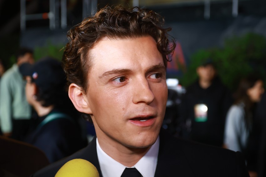 Tom Holland in a dark suit being interviewed at a press event, with a yellow microphone visible in the foreground and a blurred crowd behind him.