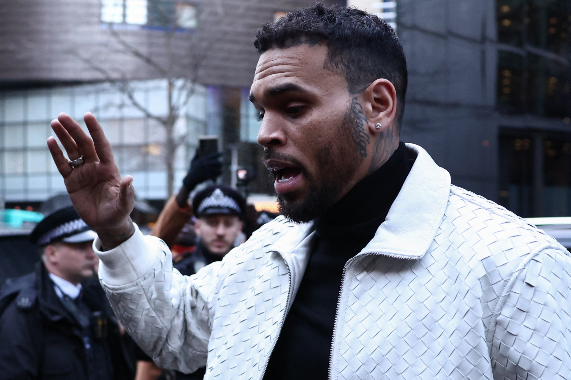Chris Brown in a white woven leather jacket raises one hand outside Southwark Crown Court, with police officers standing behind him.