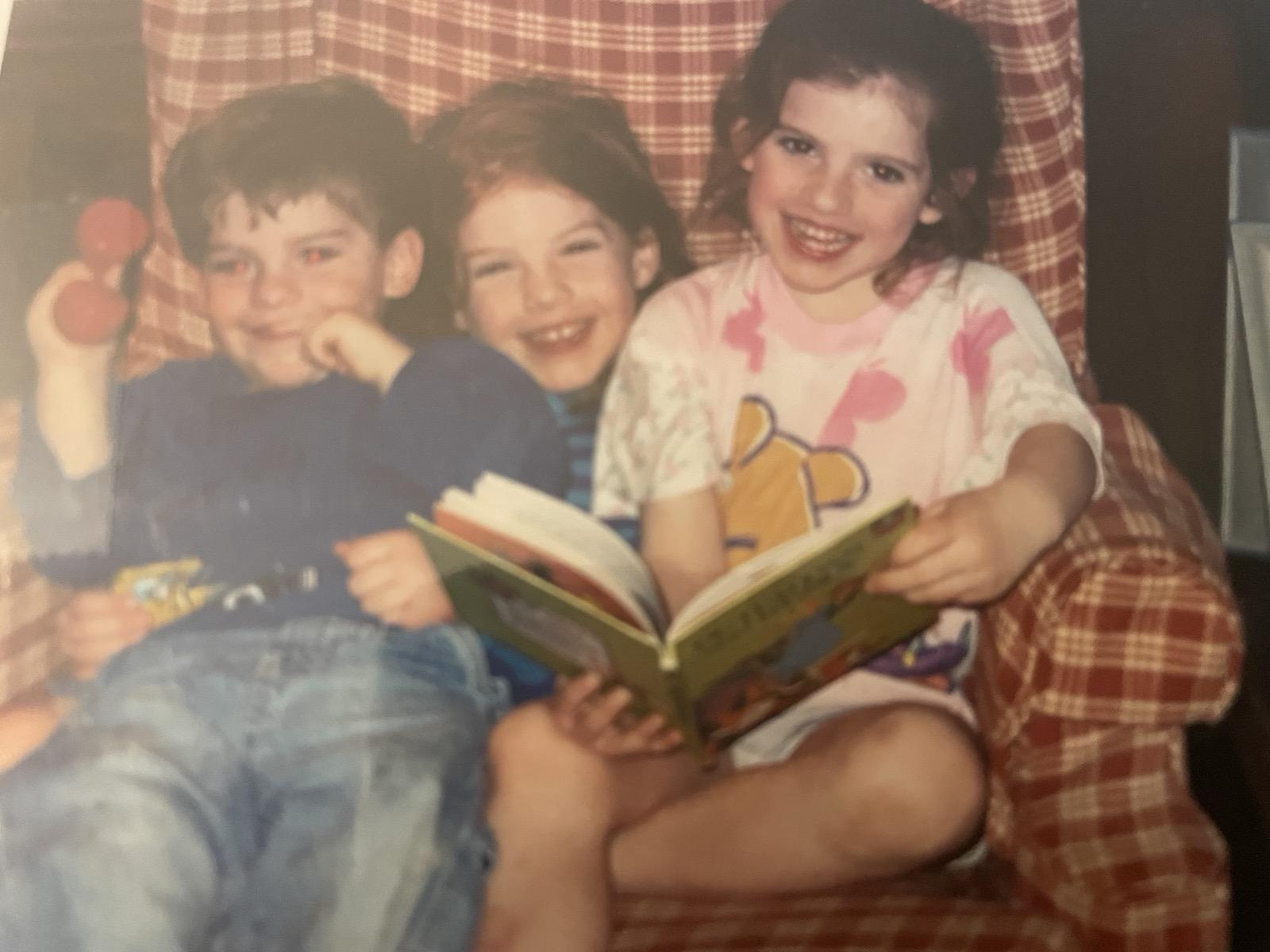 Three children — two boys and a girl — sitting together on a plaid surface, smiling and reading an open book with a green cover.