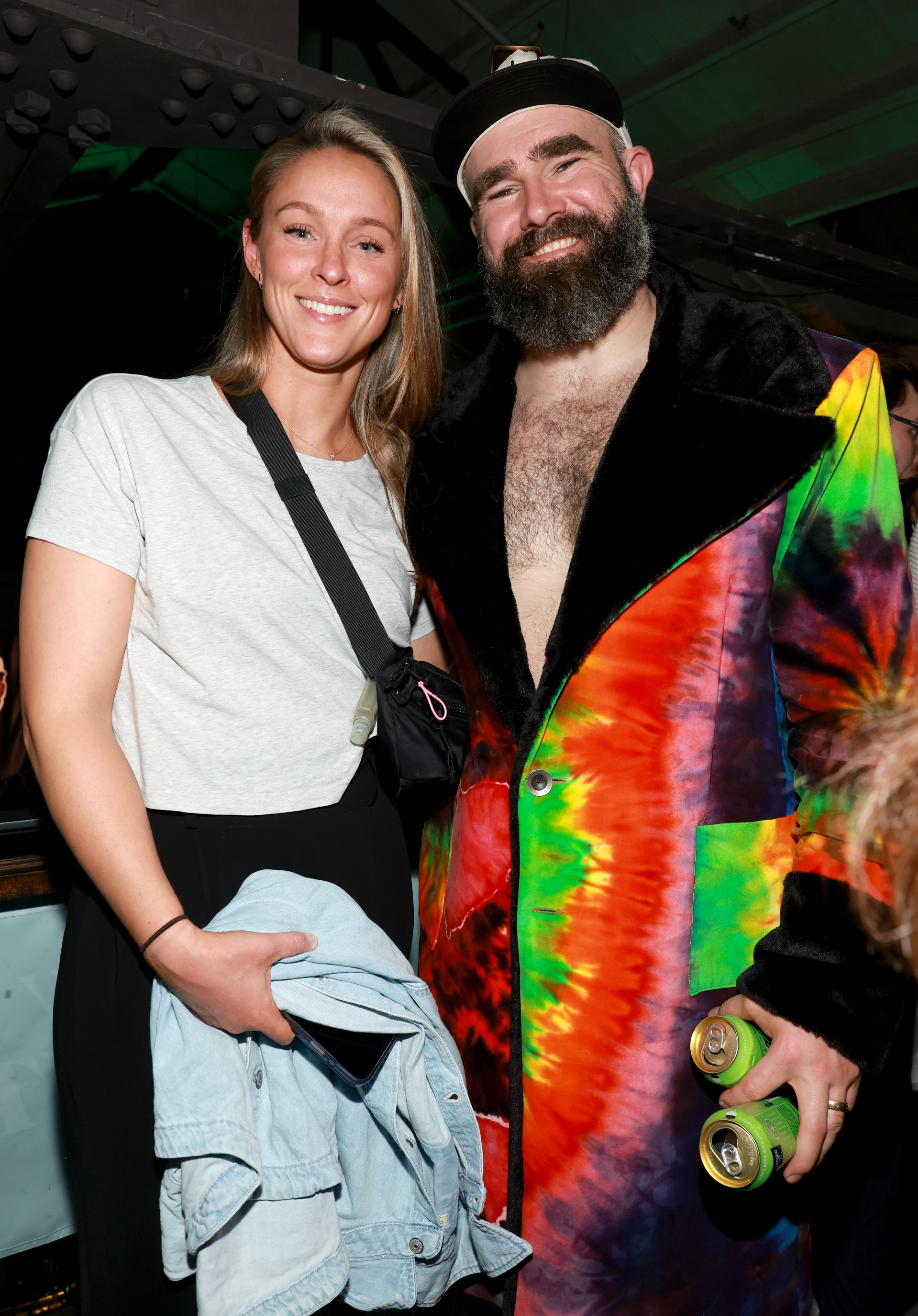 Kylie Kelce in a white crop top and Jason Kelce in a colorful tie-dye coat holding two cans, posing together at an indoor party.